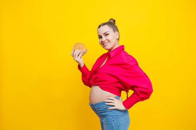 Photo funny pregnant young woman eating burger pregnant woman holding tasty sandwich while standing isolated on yellow studio wall junk food concept fast food during pregnancy