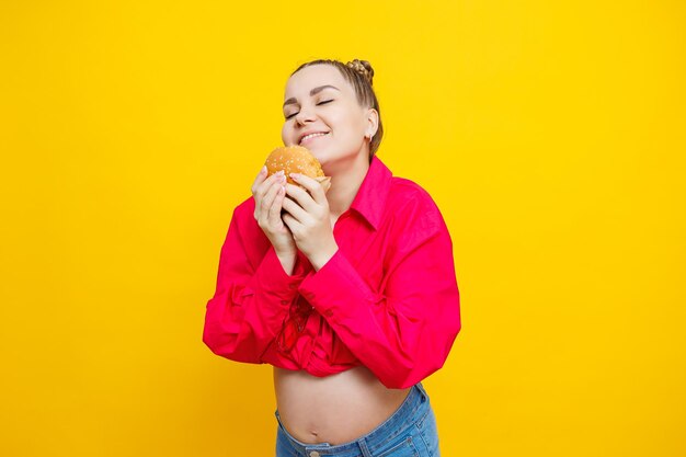 Funny pregnant young woman eating burger Pregnant woman holding tasty sandwich while standing isolated on yellow studio wall Junk food concept Fast food during pregnancy