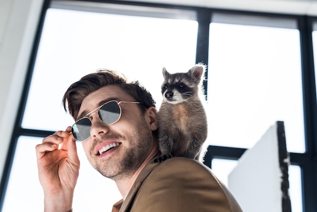 Funny raccoon sitting on shoulder of handsome smiling man in sunglasses