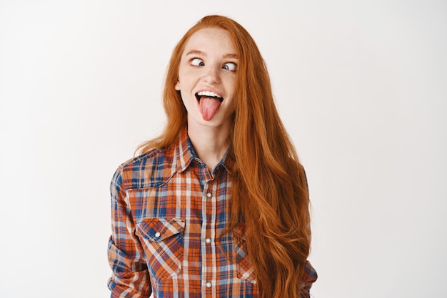 Funny redhead girl showing tongue and squinting making playful faces standing over white background