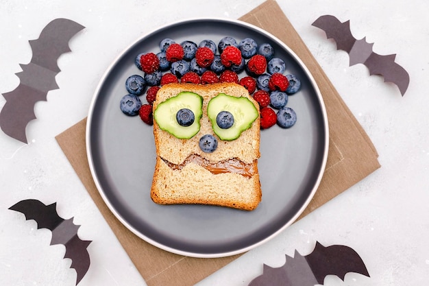 Funny scary monster face smile on halloween sandwich toast bread with peanut butter blueberry raspberry on platebats on gray background close up Kids child sweet dessert breakfast lunch food