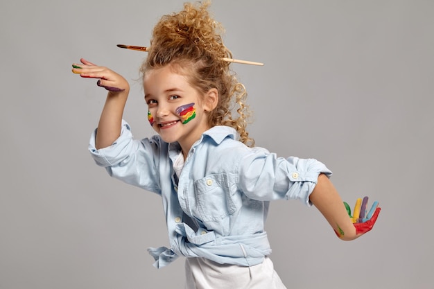 Funny schoolgirl having a brush in her chic curly blond hair, wearing in a blue shirt and white t-shirt. She is posing with a painted hands and cheeks, smiling and looking at the camera, on a gray bac