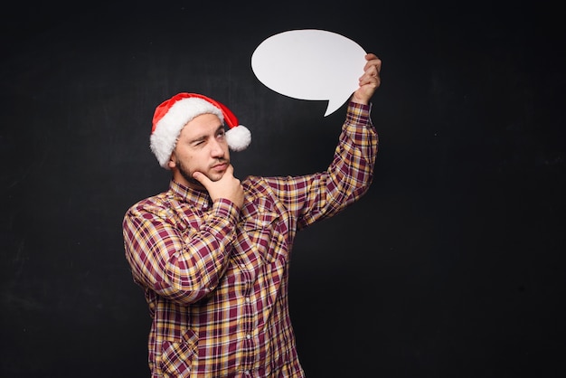 Funny serious  man in red Christmas Santa hat holds empty white cardboard as blank or mock up with copy space for text. Black background