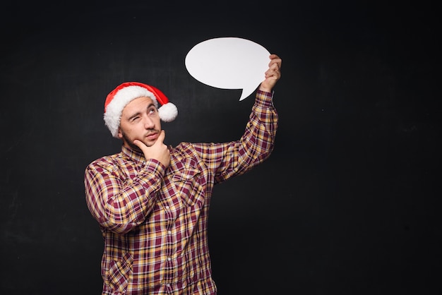 Funny serious  man in red Christmas Santa hat holds empty white cardboard as blank or mock up with copy space for text. Black background