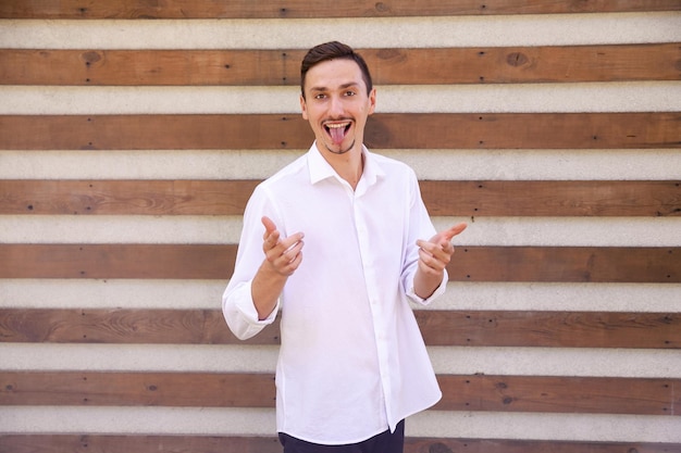 Funny smiling bearded man in a white shirt showing his tongue and pointing fingers at the camera