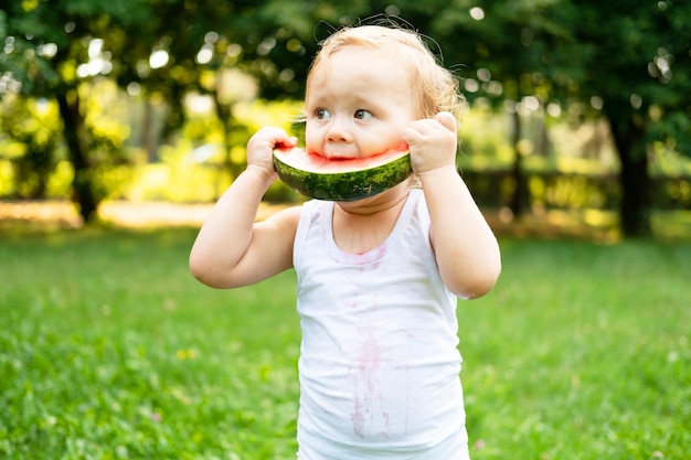 funny smiling kid boy in white bodysuit eating watermelon at green lawn