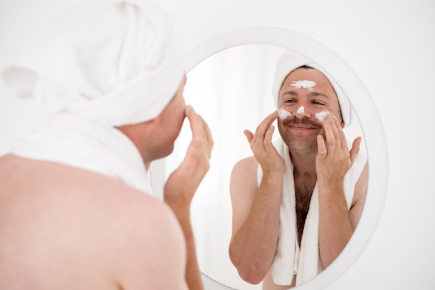 Funny smiling young man applying facial cream looking at the mirror s