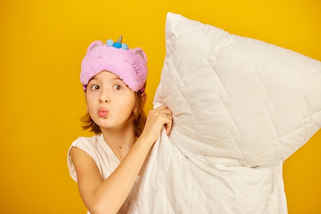 Funny teenage girl in white pyjamas with a violet sleeping mask hold pillow