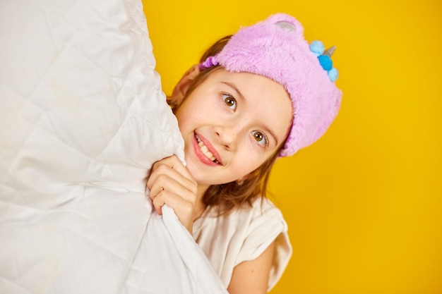 Funny teenage girl in white pyjamas with a violet sleeping mask hold pillow