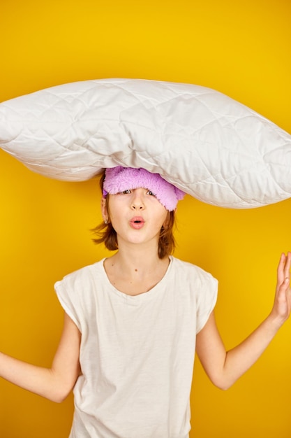 Funny teenage girl in white pyjamas with a violet sleeping mask hold pillow