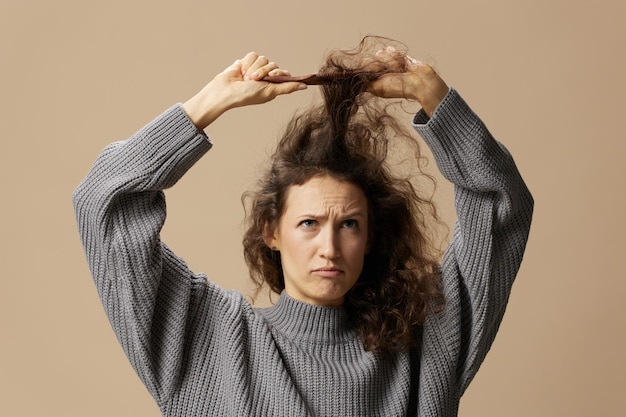 Photo funny thoughtful curly beautiful female in gray casual sweater with hairbrush comb pulls curls up posing isolated on over beige pastel background problematic unruly damaged hair concept copy space