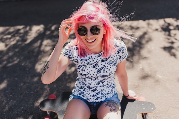 Funny top view of sporty beautiful smiling woman with blowing pink hair wearing black sunglasses with tattoo sitting on her long board waiting her friends in the park Lifestyle and sport concept