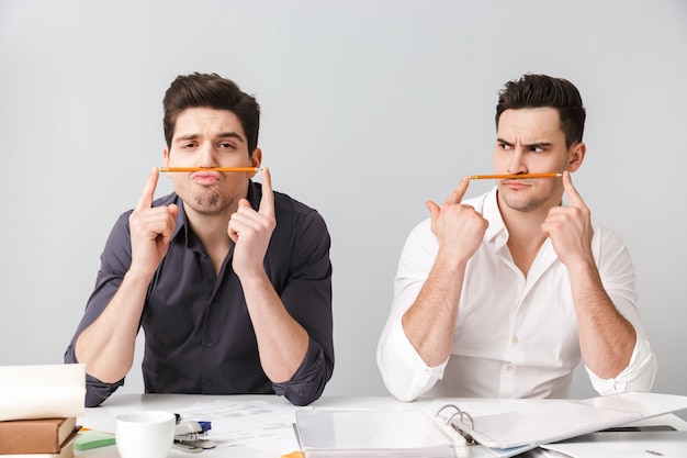 Funny two young business men have fun over grey wall.