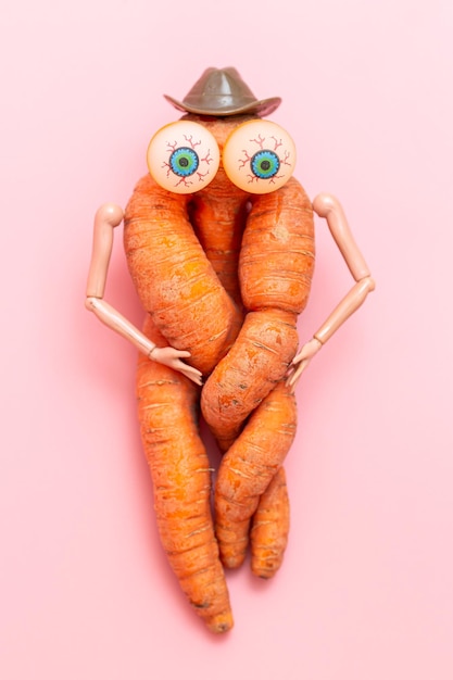 Funny ugly twisted carrot on a pink background. Carrots with hands, eyes and a hat. Vegetable toy.