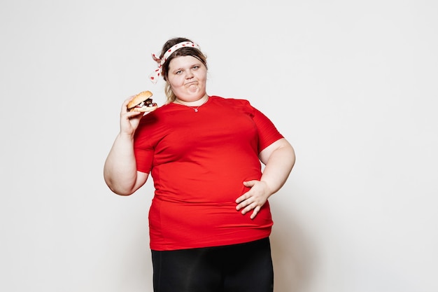 funny woman dressed in sportswear and with a bandage on her head is standing with a burger in her hand against a white wall