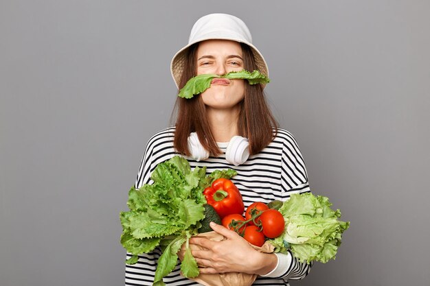 Funny woman holding vegetables isolated over gray background making mustache with green lettuce looking at camera having fun grimacing