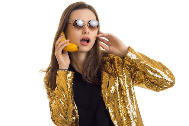 Funny woman in round sunglasses waers a golden jacket and talking in banana like a mobile phone isolated on white background