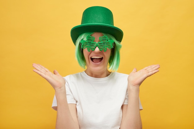 Funny woman with green hair wearing leprechaun hat and shamrock glasses posing isolated over yellow background spreading her face exclaiming with excitement