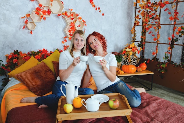 Funny women, friends, sisters sitting at cozy bed with cups, drinking tea in sweaters in fall day