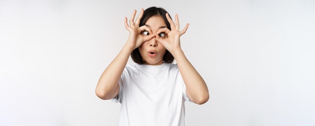 Funny young asian woman korean girl making eyes glasses gesture looking happy at camera standing over white background