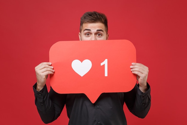 Funny young bearded guy 20s in classic black shirt posing isolated on red background. People lifestyle concept. Mock up copy space. Covering face with huge like sign from social network heart form.