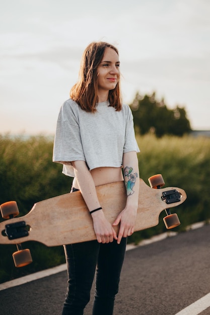 Funny young girl in jeans and top stands with big longboard green nature background high quality photo