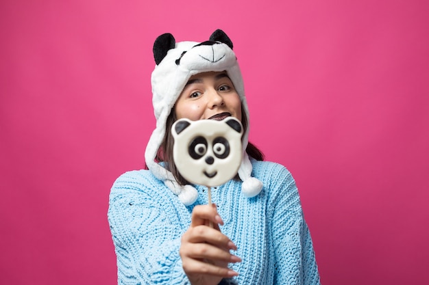 Photo funny young girl standing with yummy pandalollipop in her hand and a hat on his head on a pink