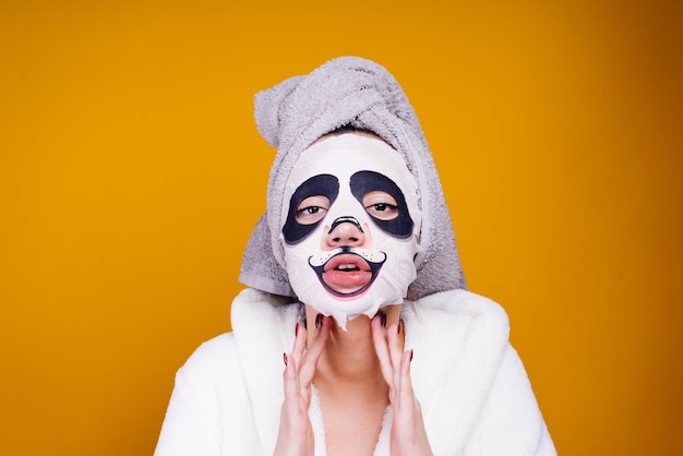 A funny young girl in a white dressing gown and with a towel on her head looks after herself on her face a moisturizing mask with a panda face