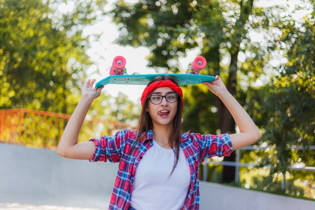 Funny young hipster woman dressed in stylish clothes holds skateboard in her hand in skatepark at bright sunny day. Summer fun