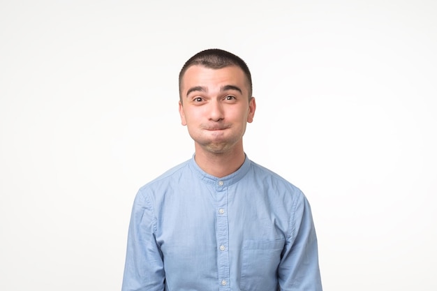 Funny young man in blue shirt grimacing staring at camera nflating cheeks holding his breath