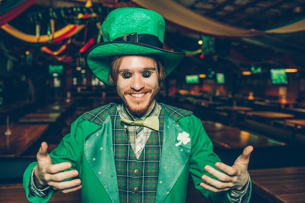 Funny young man holding coins on eyes and smile. He stand in pub alone. Guy looks happy.