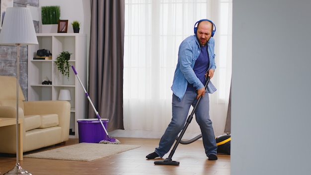 Funny young man listening music on headphone while cleaning the floor with vacuum cleaner