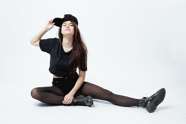 Funny young stylish girl dressed in a black top, shorts, tights and cap sits on the floor on the white background in the studio .