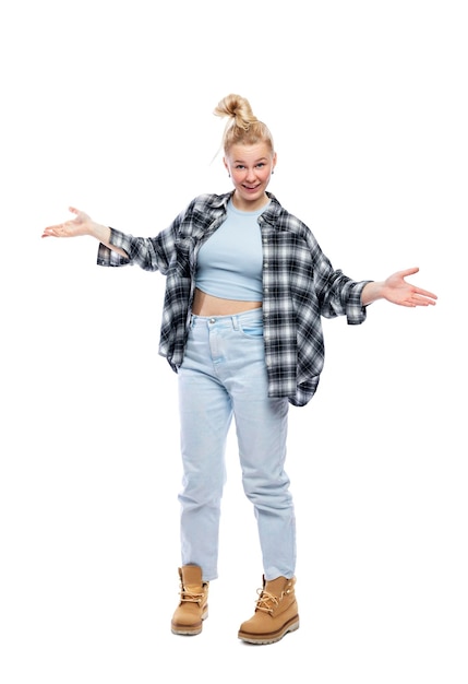 A funny young teenage girl stands with her arms spread to the sides Laughing blonde girl in jeans blue top and plaid shirt Isolated on a white background Vertical Full height