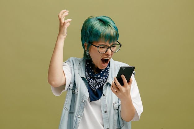 Furious young woman wearing glasses bandana on neck holding mobile phone looking at it raising hand up shouting out loudly isolated on olive green background