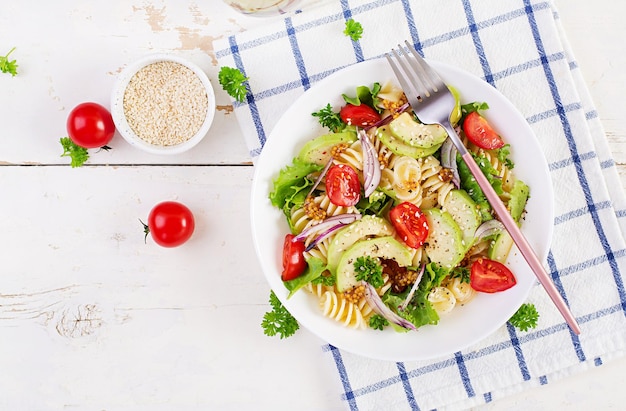 Fusilli pasta salad with avocado tomatoes fresh green lettuce red onion and mustard dressing on white background Vegetarian healthy lunch Top view flat lay