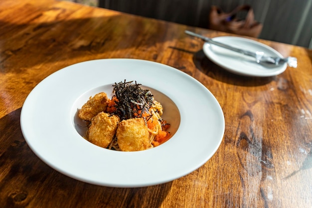 Fusion spaghetti topped with ebiko and seaweed Attached with fried breaded meat in a white round plate on a wooden table There was sunlight shining through the window Selective focus