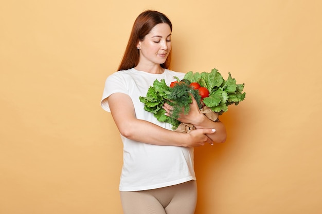 Future mother's nutrition Green market for health Adult brunette attractive pregnant woman wearing white shirt holding fresh vegetables posing isolated over beige background