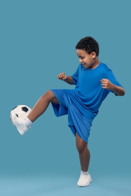 Game, football. Dark skinned tense involved boy in blue sportswear moving with soccer ball on light blue background