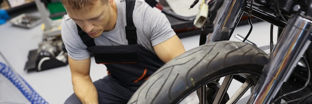 In the garage a man in overalls sits near the wheel of a motorcycle closeup checking the air