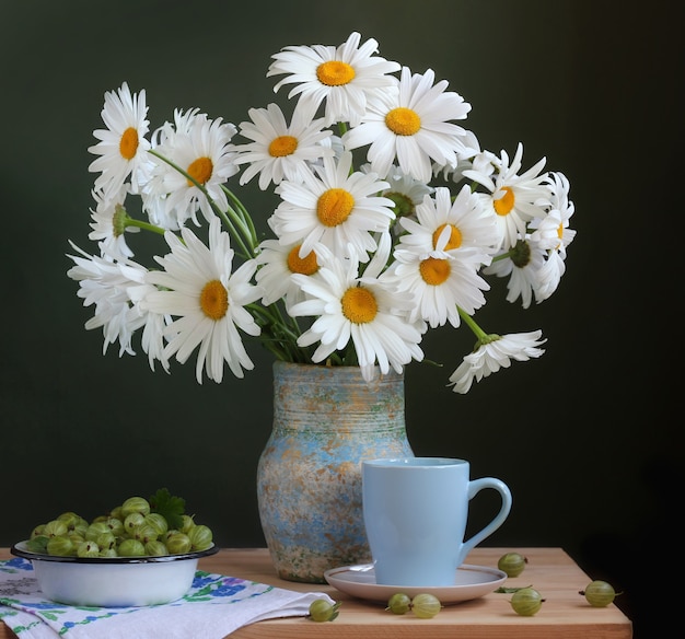 Garden chamomile in a jug on a wooden table and green gooseberry.