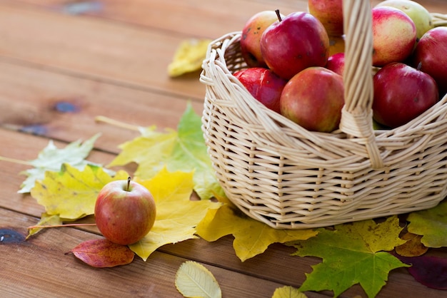 gardening, season, autumn and fruits concept - close up of wicker basket with ripe red apples and leaves on wooden table