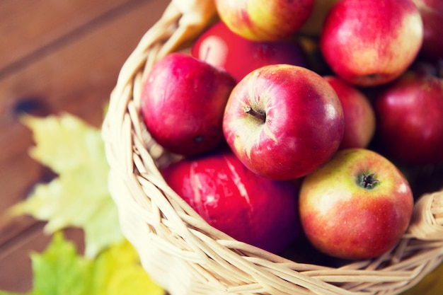 gardening, season, autumn and fruits concept - close up of wicker basket with ripe red apples and leaves on wooden table