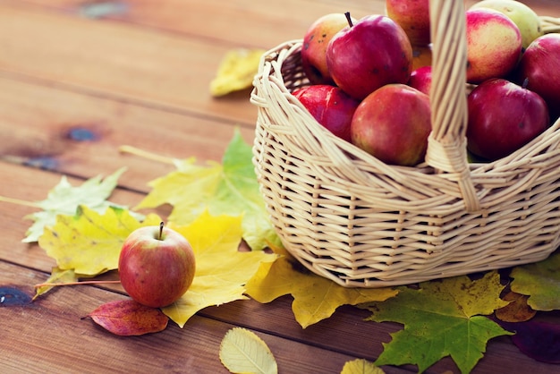 gardening, season, autumn and fruits concept - close up of wicker basket with ripe red apples and leaves on wooden table