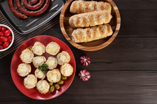 Garlic bread on a red plate on the barbecue table with sausage, cheese, rosemary, olives and cherry tomatoes. Top view.