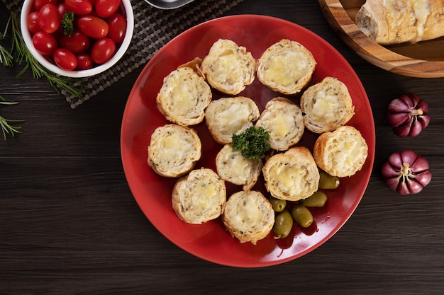 Garlic bread on a red plate on the barbecue table with sausage, cheese, rosemary, olives and cherry tomatoes. Top view.