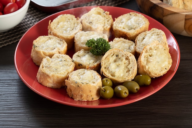 Garlic bread on a red plate on the barbecue table with sausage, cheese, rosemary, olives and cherry tomatoes.