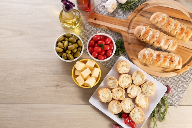 Garlic bread in white square plate on the table with cheese, rosemary, olives and cherry tomatoes. Top view.