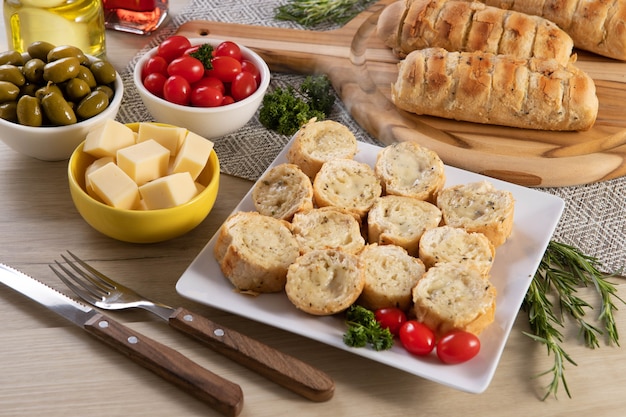 Garlic bread in white square plate on the table with cheese, rosemary, olives and cherry tomatoes.