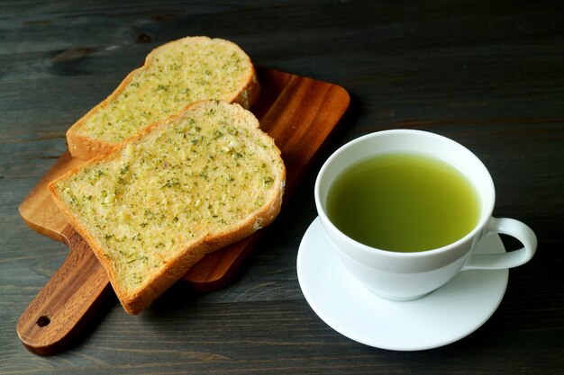 Garlic Butter Toasts on Breadboard with a Cup of Hot Green Tea on Black Wooden Table